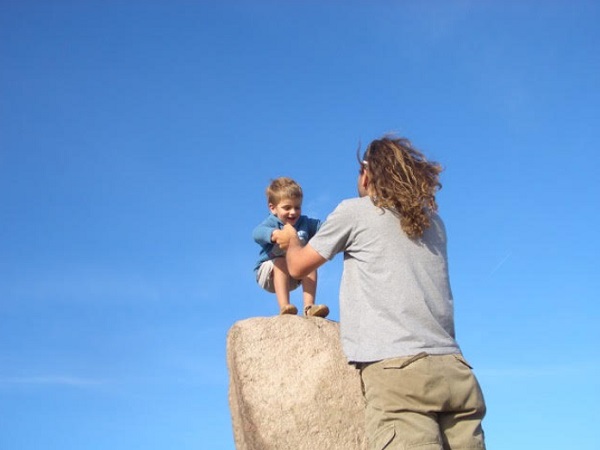 Kind sitzt auf großen Stein und Vater streckt ihn die Hände