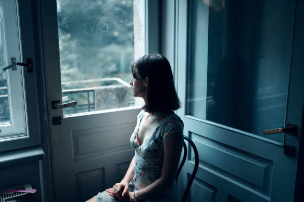 Eine Frau sitzt auf einem Stuhl und schaut durch die Glastür nach außen. Bild von Masha Raymer, Pexels.